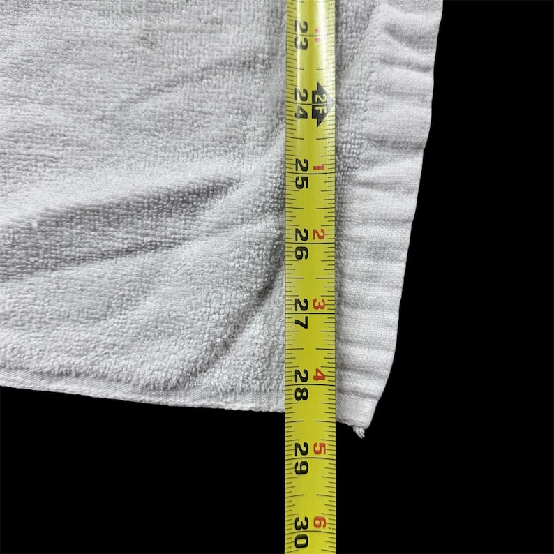 Op de afbeelding: Een lichtgrijze handdoek met een getextureerd oppervlak. Een gele meetlint is verticaal langs de rand van de handdoek geplaatst en toont metingen van 58,4 cm tot 76,2 cm.