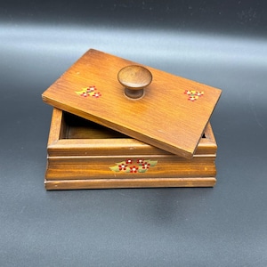 May include: A rectangular wooden box with a removable lid featuring a central knob. The lid and the front side of the box are decorated with small, painted floral motifs in red, yellow, and green. The wood has a medium brown finish.