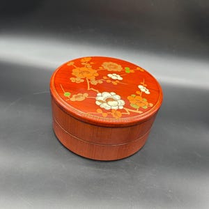 Puede incluir: Una caja redonda de madera con un acabado rojo y un diseño floral. La tapa está decorada con flores blancas y naranjas en una rama con hojas verdes.