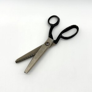 May include: Pinking shears with silver-toned metal blades and black handles. The blades feature a serrated, zigzag edge design for cutting fabric. The two blades are joined by a central metal screw, and the handles have a worn, textured finish.