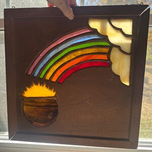 May include: Stained glass panel featuring a rainbow with red, orange, yellow, green, blue, and pink arches. A pot of gold sits below the rainbow. The panel is framed in a dark brown wooden frame.