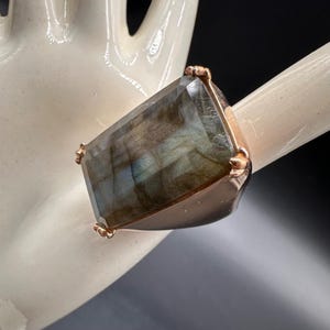 May include: A ring featuring a large, rectangular, faceted labradorite gemstone with grey and blue iridescent tones. The stone is set in a polished rose gold-toned metal band, secured by four small prongs at the corners of the gemstone setting.