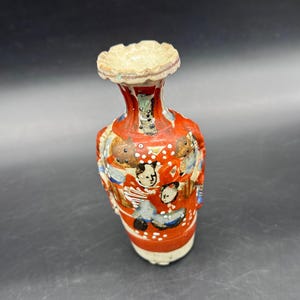 May include: A small, antique ceramic vase with a flared rim and a red body. The vase is decorated with painted figures and white polka dots. The rim is a light beige color. The vase is approximately 6 inches tall.