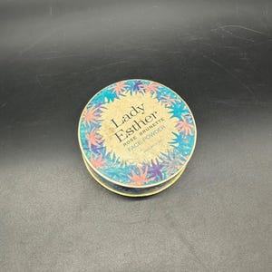 May include: A round, blue and pink compact with a floral design and the text "Lady Esther Rose Brunette Face Powder" printed on the lid.