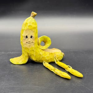 May include: A yellow carved wooden banana figurine with a smiling face, textured surface, and a curled top section. The figure features two dangling yellow legs attached by thin cords, extending outward from the base of the banana body.