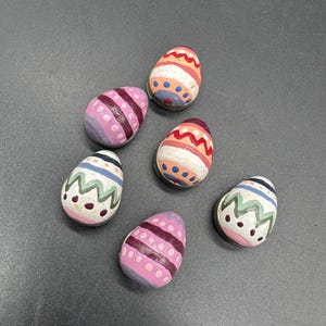 May include: Six hand-painted wooden Easter eggs in various colors. Each egg features unique designs, including stripes, dots, and zigzags. The color palette includes pink, purple, orange, blue, and white, creating a festive and decorative look.