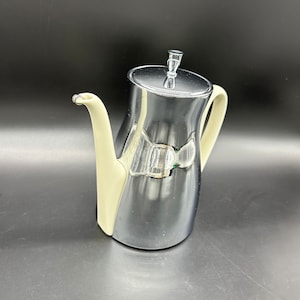 Puede incluir: Una tetera plateada con un asa y un pico blancos. La tetera tiene una superficie brillante y reflectante.