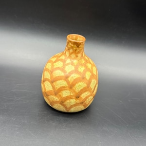 May include: A small, round ceramic vase with a narrow neck. The vase features a yellow and orange pattern of overlapping, curved shapes. This decorative vase is a handcrafted item, suitable for home decor.