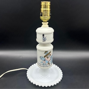 May include: A vintage white table lamp with a brass bulb socket. The lamp features a stacked design with cartoon illustrations on the cylindrical shades. It sits on a white, scalloped base and has a white cord.