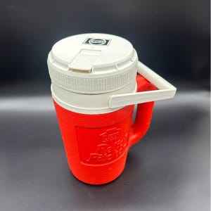 Puede incluir: Una nevera Pepsi roja y blanca vintage con asa y tapa blancas. La tapa presenta el logotipo de Pepsi. La nevera tiene un cuerpo rojo y una parte superior blanca. La nevera está diseñada para mantener las bebidas frías.