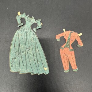 Puede incluir: Dos conjuntos de mu&ntilde;ecas de papel vintage. Uno es un vestido color turquesa con detalles florales. El otro es un traje pantal&oacute;n rojo con ribetes verdes. Ambos conjuntos tienen pesta&ntilde;as para sujetarlos a una mu&ntilde;eca de papel.