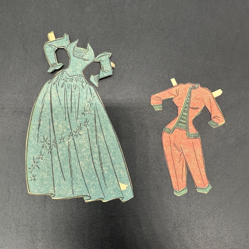 Puede incluir: Dos conjuntos de mu&ntilde;ecas de papel vintage. Uno es un vestido color turquesa con detalles florales. El otro es un traje pantal&oacute;n rojo con ribetes verdes. Ambos conjuntos tienen pesta&ntilde;as para sujetarlos a una mu&ntilde;eca de papel.