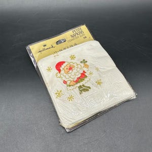May include: A package of Hallmark Petite Napkins, coaster size, featuring a Santa Claus design. The napkins are white with a repeating pattern of gold snowflakes. The package dimensions are 3.25 inches by 3.25 inches.