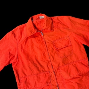 May include: Bright orange jacket with a collared neckline, a full-length zipper, and multiple front pockets. The jacket appears to be made of a lightweight material, suitable for layering. The garment is a solid, vibrant color.