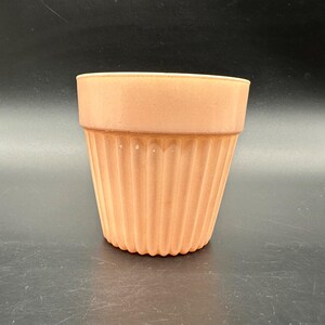 May include: A peach-colored ceramic planter with a ribbed exterior. The top of the planter has a smooth, untextured band. The planter is designed to hold plants and flowers. The background is black.