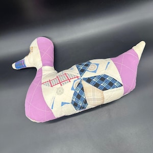 May include: A handmade duck-shaped pillow with a patchwork design. The duck features a lavender head and tail, with a body composed of various fabric patterns in blue, white, and tan. The pillow is a decorative item.
