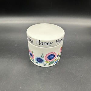 May include: White ceramic honey pot with a floral design. The lid has the words "Honig Honey Honig" written around the edge.