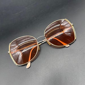 Könnte beinhalten: Quadratische Sonnenbrille mit braunen Gläsern und goldfarbenen Metalldetails. Die Fassungen haben ein Retro-Design mit einem dünnen silbernen Steg und orangefarbenen Akzenten. Die Sonnenbrille ist vor einem dunklen Hintergrund platziert.