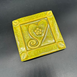 May include: A square ceramic coaster in a yellow-green hue, featuring a heart and paw print design. It has a raised border and leaf details. The heart and paw print are a lighter shade of yellow. The coaster is approximately 10 cm square.
