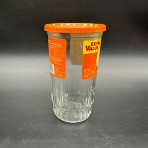 May include: A clear glass jar with an orange lid. The jar has a ridged design on the bottom half and a label that reads "EXTRA VALUE" in yellow and red. The label also indicates a net weight of 16 oz (1 lb) or 453g.