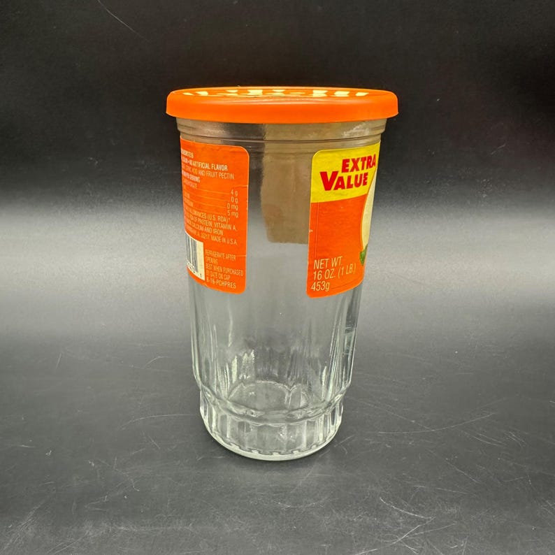 May include: A clear glass jar with an orange lid. The jar has a ridged design on the bottom half and a label that reads "EXTRA VALUE" in yellow and red. The label also indicates a net weight of 16 oz (1 lb) or 453g.