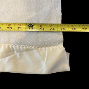 May include: A white blanket with a cream-colored satin trim. A yellow measuring tape shows the blanket's width is approximately 70 to 77 inches.