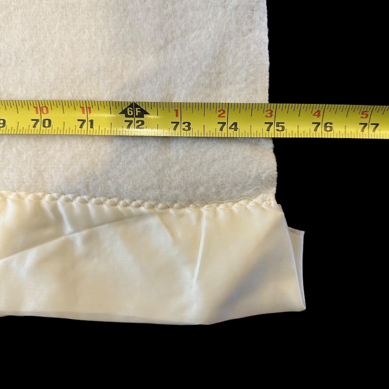 May include: A white blanket with a cream-colored satin trim. A yellow measuring tape shows the blanket's width is approximately 70 to 77 inches.