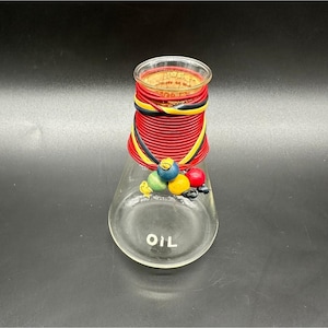 Könnte beinhalten: Eine klare Ölflasche aus Glas mit einem dekorativen roten, schwarzen und gelben Deckel. Die Flasche hat einen bauchigen Boden und das Wort "OIL" ist auf die Vorderseite gedruckt. Bunte, runde Verzierungen sind am Boden befestigt.