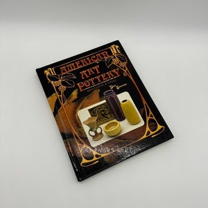 May include: A black hardcover book titled "American Art Pottery: A Collector's Guide to Identification & Values" by Dick Sutter. The book cover features a decorative border with images of pottery.