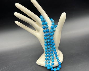 May include: A long necklace of bright blue beads draped over a white ceramic hand. The beads are a vibrant turquoise color, and the necklace appears to be made of multiple strands. The ceramic hand is smooth and glossy, with a neutral color.