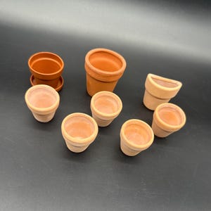 Puede incluir: Una colección de pequeñas macetas de terracota de varios tamaños y formas. Algunas macetas son cilíndricas, mientras que otras tienen un diseño más redondeado o rectangular. Un juego incluye un platillo a juego. Estas macetas son adecuadas para plantas pequeñas.