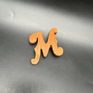May include: A wooden letter "M" in a warm, reddish-brown hue. The handcrafted initial has a decorative, cursive style with rounded edges. The wooden letter is likely for home decor or craft projects.