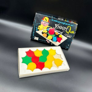 May include: A vintage jigsaw puzzle game called "K-Wazy Quilt" with a colourful illustration of a person wearing a hat and holding a piece of the puzzle. The box is black with white text and the puzzle pieces are on a white surface.