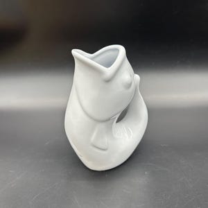 May include: A white ceramic vase in the shape of a fish. The vase has a wide opening at the top and a curved tail. The fish's mouth is open, and its body is smooth and glossy. The vase is a decorative item.