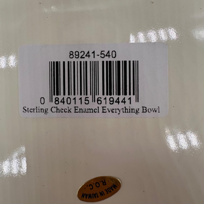 Op de afbeelding: Een wit rechthoekig productlabel met de tekst 89241-540, een streepjescode en de tekst Sterling Check Enamel Everything Bowl. Onder het label bevindt zich een kleine ovale gouden sticker met de tekst MADE IN TAIWAN R.O.C. op een glanzend oppervlak.