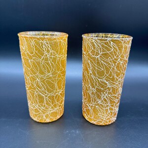 May include: Two cylindrical drinking glasses featuring a textured amber-colored glass surface overlaid with a white, chaotic, string-like pattern. The glasses have a straight-sided design with a consistent diameter from the base to the rim.