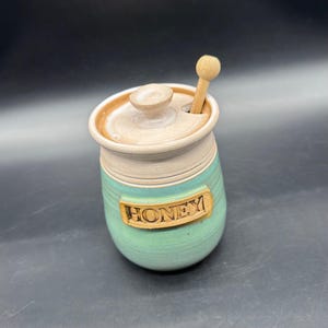 May include: A ceramic honey pot with a lid and a wooden honey dipper. The pot is light green with a beige top and a brown rim. The word "HONEY" is embossed on the front.