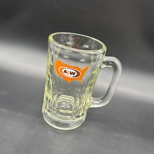 May include: Clear glass mug with a handle. The mug has a textured surface and features an orange and brown logo with the letters "A&W" inside a map of the United States. The mug is designed for beverages.