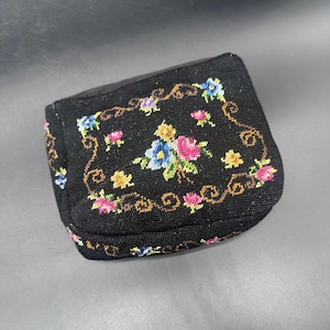 May include: A small, black, vintage purse with a floral needlepoint design. The needlepoint features pink, blue, and yellow flowers with green leaves, framed by a brown, scrolling border. The purse has a rounded shape and a black fabric top.