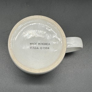 May include: White ceramic mug with a brown stripe around the rim. The bottom of the mug has the text "MADE IN KOREA F.T.D.A. &copy;1984" printed in black.