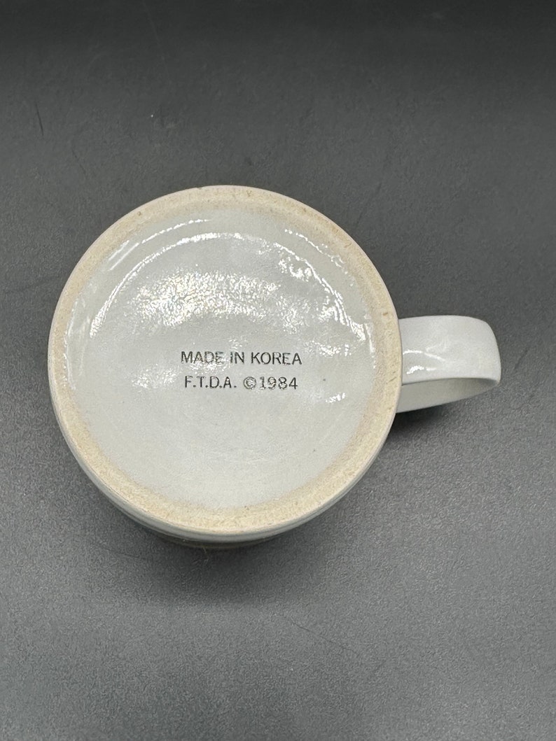 May include: White ceramic mug with a brown stripe around the rim. The bottom of the mug has the text "MADE IN KOREA F.T.D.A. &copy;1984" printed in black.