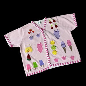 May include: A pink short-sleeved cardigan with a white stripe down the front. The cardigan is decorated with embroidered fruit, ice cream, and swimsuit designs.