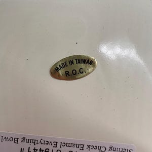 Op de afbeelding: Een ovale goudkleurige sticker met zwarte tekst MADE IN TAIWAN R.O.C. bevestigd op een glad wit oppervlak. Onder de sticker is een gedeeltelijk wit label zichtbaar met de zwarte tekst Sterling Check Enamel Everything Bowl.