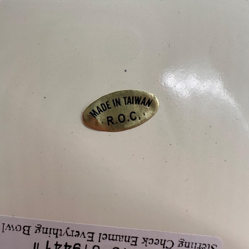 Op de afbeelding: Een ovale goudkleurige sticker met zwarte tekst MADE IN TAIWAN R.O.C. bevestigd op een glad wit oppervlak. Onder de sticker is een gedeeltelijk wit label zichtbaar met de zwarte tekst Sterling Check Enamel Everything Bowl.