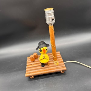 Puede incluir: Una lámpara de mesa decorativa con una figura de pájaro amarillo que lleva un sombrero de pirata negro y un parche en el ojo, sobre una base de madera en forma de balsa con dos barriles. Un poste de madera sostiene un casquillo metálico.