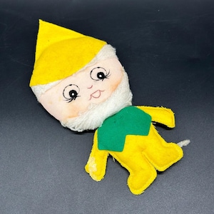 May include: A vintage felt doll with a yellow hat and body, a green collar, and a white face and neck. The doll has a smiling face with black eyes and a red mouth. The doll is lying on a dark surface.