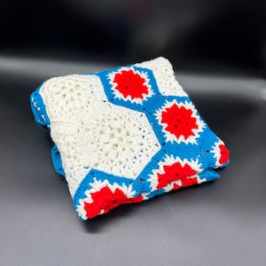 Puede incluir: Una manta de ganchillo blanca con un patrón geométrico de flores rojas y azules. La manta está doblada por la mitad y tiene una superficie suave y texturizada.