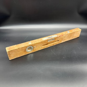 May include: A rectangular wooden spirit level tool featuring a natural light brown wood grain finish. The tool includes three integrated glass vials filled with liquid for horizontal and vertical leveling, set into metal frames along the top and side.