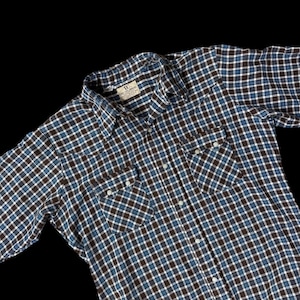 Könnte beinhalten: Ein blau-braun-weiß kariertes Button-Down-Hemd. Das Hemd hat zwei Vordertaschen mit weißen Knöpfen und einen Kragen. Das Hemd ist aus einem weichen, gewebten Stoff gefertigt. Das Lucky Brand-Etikett ist am Kragen sichtbar.