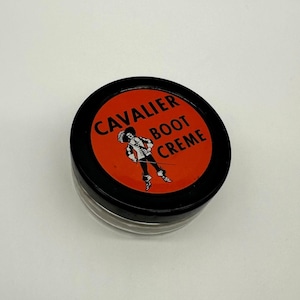 May include: A round, black container of Cavalier Boot Creme. The lid is orange with black text and a graphic of a cavalier. The container is on a white surface.